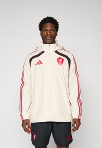White athletic jacket with red accents and black stripes, featuring a hood and zipper. Worn with dark shorts, displaying the Liverpool FC logo.