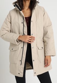 Parka acolchada beige con cierre de cremallera y botones a presión, capucha desmontable y bolsillos frontales; presenta una textura suave y aislamiento térmico.