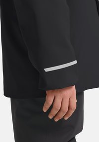 Schwarze wasserdichte Jacke mit einem Ärmel, der ein reflektierendes silbernes Detail in der Nähe des Bündchens aufweist. Die Textur ist glatt und der Schnitt ist entspannt.