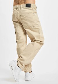 Beige cargo pants with a relaxed fit, featuring side pockets, back pockets, and a textured fabric. Worn with white athletic sneakers.