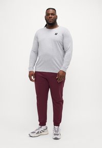 Grå langermet t-skjorte med svart logo på brystet, paret med burgunder joggebukser. Modell som har på seg grå og hvite joggesko.