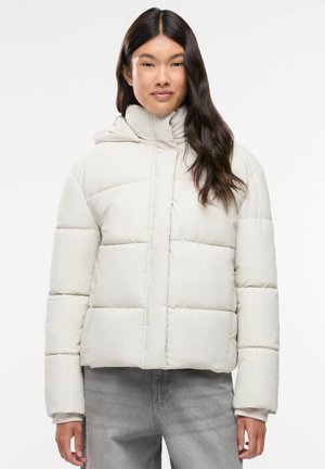 Donna con capelli lunghi e scuri che indossa una giacca imbottita beige chiaro, in piedi davanti a uno sfondo bianco semplice.