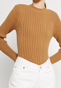 Brauner, gerippter Strickpullover mit langen Ärmeln und hohem Kragen, der vertikale Streifen und eine figurbetonte Silhouette aufweist, kombiniert mit weißen, hochgeschnittenen Jeans.