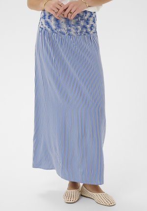 Kvinde iført en lang blå- og hvidstribet nederdel med elastisk linning, stående barfodet i beige vævede slip-on sko.