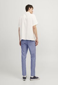 Camisa blanca de manga corta con tejido texturizado, combinada con pantalones azul claro y zapatillas oscuras, vista desde la parte trasera sobre un fondo neutro.