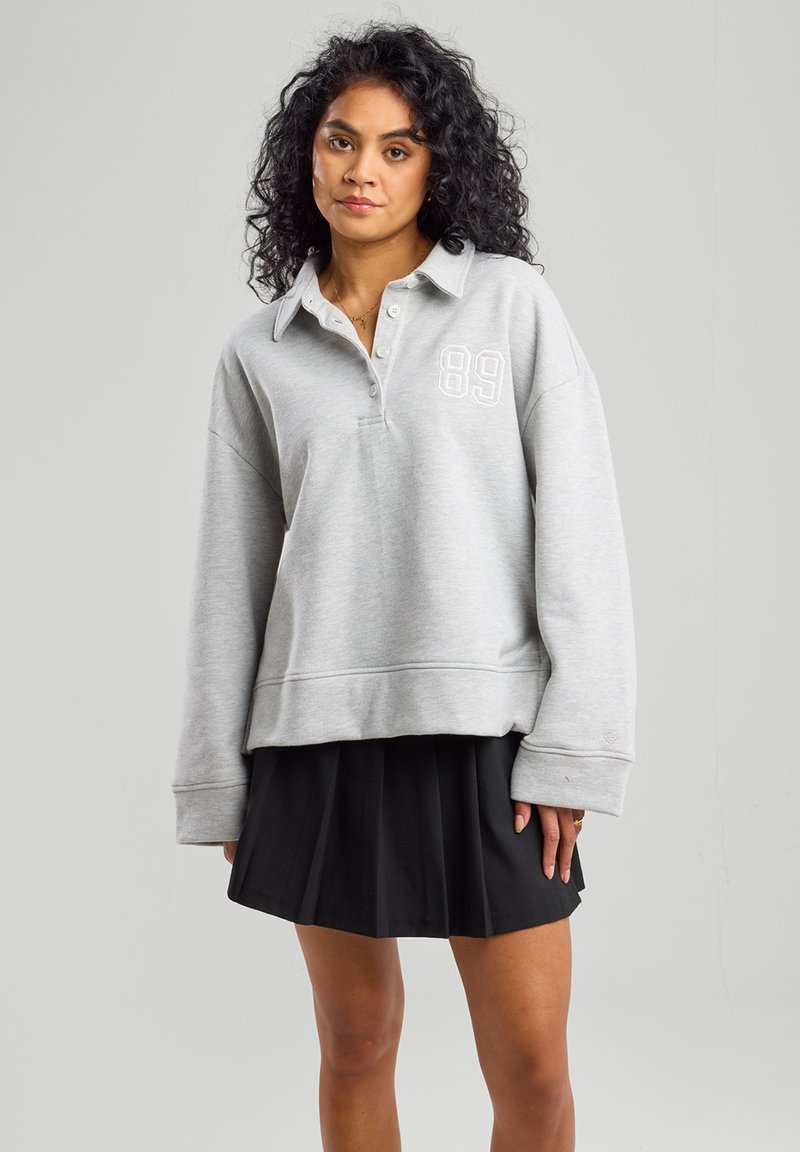 Harmaa pulloversweater, jossa on kaulus, kolme nappia ja valkoinen "89" -painatus. Rentoa istuvuutta ja ribbauskudin hihansuissa, yhdistetty mustan pliseeratun hameen kanssa.