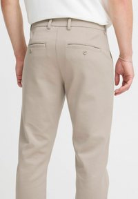 Pantalones beige con una textura suave, que cuentan con dos bolsillos traseros, cada uno con cierre de botón. Ajuste entallado con un diseño de pierna recta.