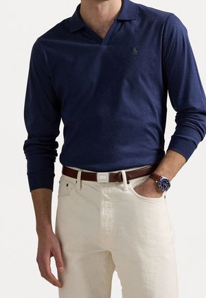 Mand iført navyblå langærmet poloshirt, beige bukser med brun bælte og brun læderarmbåndsur med blå urskive, hånd i lommen.