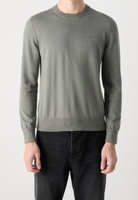 Mies, który ma na sobie jasnoszary sweter z długim rękawem z małym logo "BOSS" na piersi, zestawiony z czarnymi spodniami, stoi na jednolitym tle.