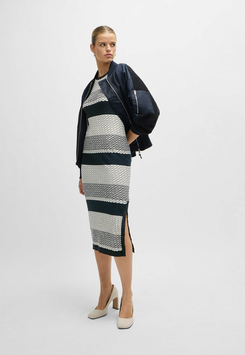 Figurbetontes Kleid mit marineblauen, weißen und grünen Streifen; strukturierte Musterung. Ergänzt durch eine schwarze Bomberjacke und neutrale Blockabsatzschuhe.