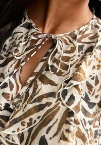 Djurmönstrad blus i lätt, genomskinligt tyg med volangdetaljer och en nyckelhålsringning som knyts med en rosett. Färgerna inkluderar beige, brunt och svart.