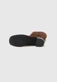 Stivale alla caviglia in suede marrone con punta tonda, suola in gomma nera e dettaglio di branding metallico sul lato. Texture liscia in tutta la superficie.