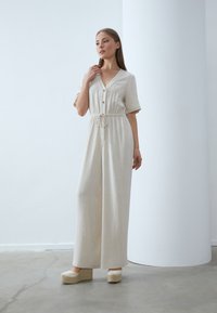 Beige linne jumpsuit med v-ringning, korta ärmar, elastisk midja och träknappar. Matchad med beige plattform espadriller. Minimalistisk design.