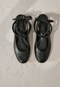 Mustad nahast balletitaldsed, ümarate varvastega ja nööri detailiga pahkluupotis. Sile tekstuur ja minimalistlik disain.