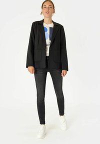 Črn krojeni blazer z dvema sprednjima žepoma, v kombinaciji z belo grafično majico, črnimi kavbojkami in belimi športnimi čevlji. Gladek material, strukturiran dizajn.