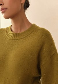 Frau in einem senfgelben, gesprenkelten Strickpullover mit geripptem Rundhalskragen vor einem neutralen Hintergrund.