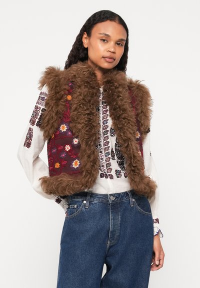 Hnedá vestu s kožušinovým límcom a kvetinovou výšivkou, kombinovanú s bielou vyšívanou košeľou a modrými džínsami. Mäkká textúra a živé vzory.