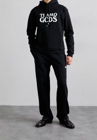 Czarna bluza z kapturem z białym napisem "TI AMO GODS", w połączeniu z czarnymi dresami. Dodatki obejmują błyszczące czarne eleganckie buty. Prosty design, miękka faktura.