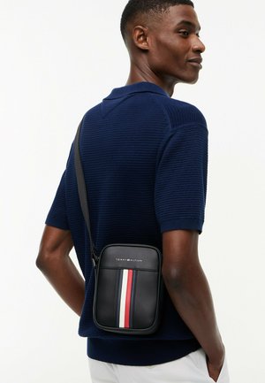 Mann trägt ein marineblaues, strukturiertes Kurzarmhemd mit schwarzer Umhängetasche mit roten, weißen und marineblauen vertikalen Streifen und Tommy Hilfiger-Logo.