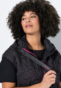 Chaleco negro acolchado con costuras rosas y cierre frontal con cremallera. Cuenta con un cuello alto y tirantes, superpuesto a una camiseta negra.