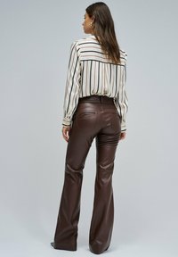 Vrouw met de rug naar de camera, gekleed in een witte blouse met lange mouwen en verticale strepen, en hoge bruine leren broeken met een flared pasvorm.