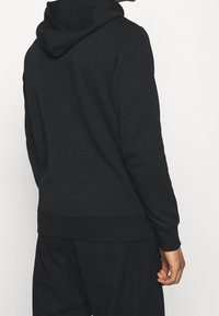 Felpa nera con cappuccio realizzata in tessuto morbido, caratterizzata da orli e polsini a coste, con una tasca a marsupio frontale e dettagli minimi.