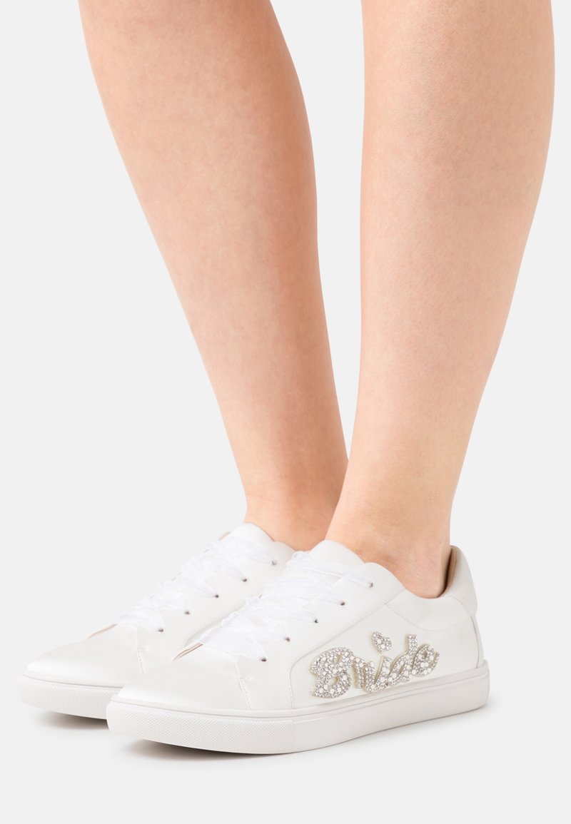 White sneaker with smooth synthetic material, lace-up design, and rhinestone "Bride" detail on the side. Low-cut ankle with textured sole.