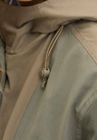 Veste légère bicolore avec une capuche beige et un corps vert doux. Comprend un cordon de serrage avec un curseur et des détails de couture subtils.