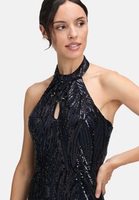 Kobieta w czarnej, bezrękawnikowej sukience z cekinami, z dekoltem w kształcie łezki, patrząca na bok z ręką na biodrze, na białym tle.