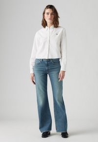 Chemise blanche à boutons avec un col, associée à un jean flare bleu clair. Présente un petit logo sur la chemise, complétée par des chaussures noires.