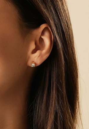 Goldener Ohrstecker mit klarem rundem Edelstein, getragen am linken Ohr einer Person mit glatten braunen Haaren.