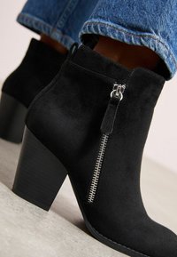 Botines de ante negros con un acabado texturizado, detalle de cremallera lateral y tacón apilado. Jeans parcialmente visibles en la parte superior.