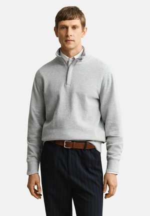 Homme portant un pull gris clair à fermeture éclair quart, par-dessus une chemise blanche et un pantalon sombre à fines rayures avec une ceinture marron, debout devant un fond blanc.