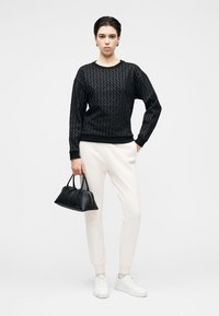 Camisola preta texturizada com padrão de diamante, calças de fato de treino creme, mala de mão preta e sapatilhas brancas, exibidos contra um fundo neutro.