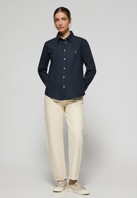 Camicia blu navy con bottoni in tessuto morbido, caratterizzata da un colletto a punta e un piccolo emblema sul petto. Abbinata a pantaloni crema e scarpe chiare.