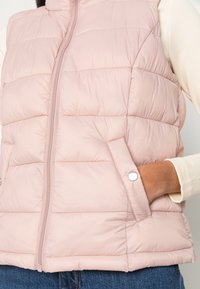Chaleco acolchado de color rosa claro con textura acolchada vertical, cremallera frontal y bolsillos laterales. Tejido suave con un ajuste ceñido. Ideal para superponer.