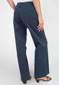 Wijde broek in donkerblauwe stof, met een achterzak en een tailleband. Vergezeld van zwarte sandalen met bandjes.