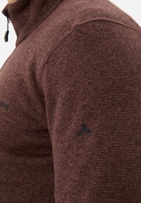 Maglione lavorato a maglia marrone con colletto alto, dotato di chiusura con zip. Tessuto con una leggera trama e un logo nero sulla manica sinistra, finitura del tessuto liscia.