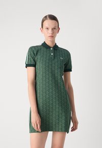 Rochie polo verde cu un model geometric repetitiv, mâneci scurte și un guler negru. Dispune de trei butoane și dungi albe pe laterale.