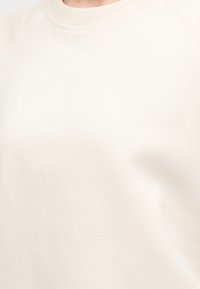 Κοντινό πλάνο ενός μπεζ πουλόβερ με στρογγυλή λαιμόκοψη, που φοριέται από άτομο, δείχνοντας την υφή του υφάσματος και τις λεπτομέρειες στη λαιμόκοψη.