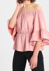 Blouse rose clair à épaules dénudées avec un col élastique froncé et des manches évasées. Tissu doux avec un détail en bas de péplum.