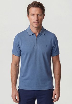 Man wearing a blue short-sleeve polo shirt with a zip collar and small logo on left chest, standing against a plain background.