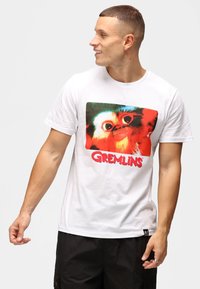 Valkoinen puuvillapaita, jossa on suuri värikäs Gremlins-kuviointi. Lyhyet hihat, rento malli, pyöreä pääntie ja musta label-aksentti.