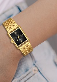 Kuldne ristkülikukujuline kell tekstuuriga musta kellaga, kahe kuldse käega ja reguleeritava kuldse lingi rihmaga, mis on kantud randmel.