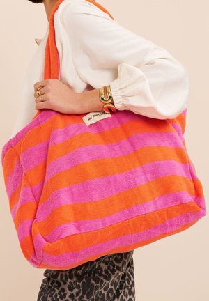 Persona che indossa una camicetta bianca e pantaloni a stampa leopardata tiene una grande borsa a righe rosa e arancioni con l'etichetta "My Jewellery" ben visibile.