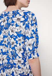 Blusa floral azul com lírios brancos contornados em preto, com um corte descontraído e mangas elásticas para um toque extra de textura e detalhe.