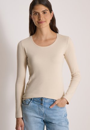 Femme aux longs cheveux foncés portant une chemise beige à manches longues et un jean bleu clair, debout avec une main dans la poche sur un fond uni.