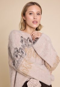 Overdimensjonert, lys beige genser med myk, fluffy tekstur. Har intrikate mørkebrune og lysebrune mønstre på ermene. Rund halslinning.