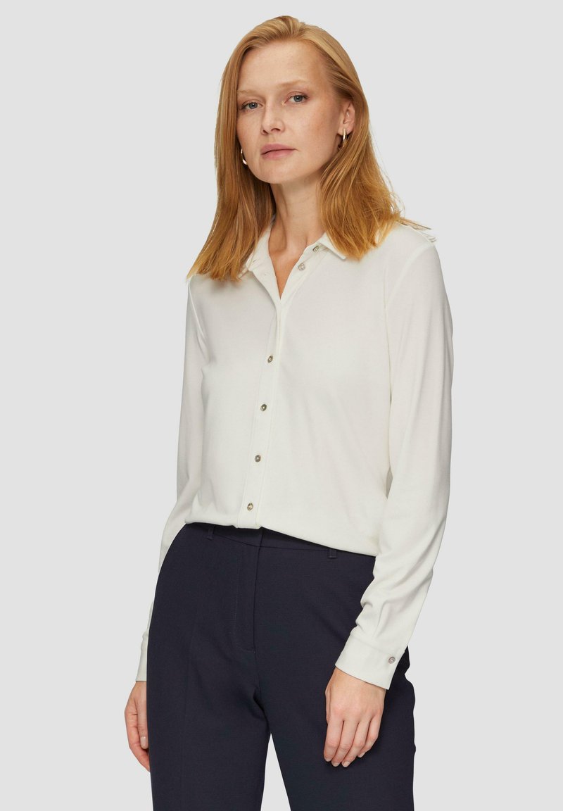 White long-sleeve blouse with a button-up front, featuring a collar and cuffs with button details. Smooth fabric; paired with navy trousers.