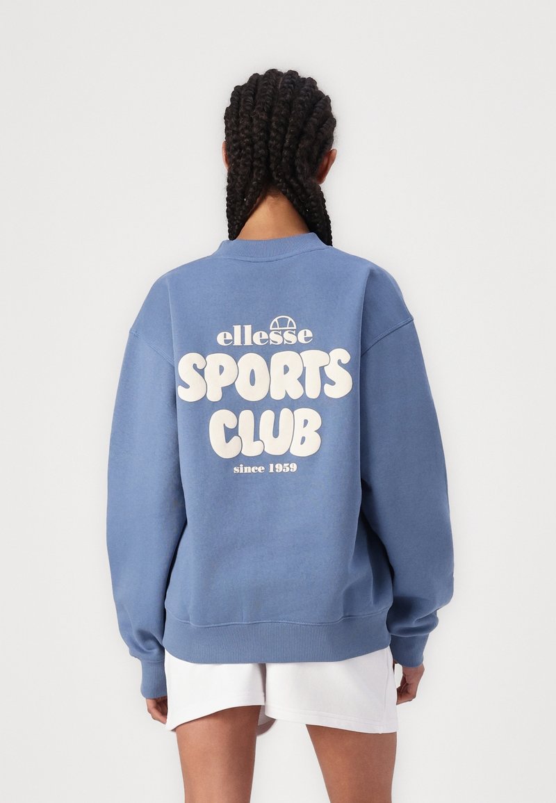 Personne aux cheveux tressés porte un sweat-shirt bleu avec l'inscription « ellesse SPORTS CLUB since 1959 » et un short blanc, debout devant un fond uni.